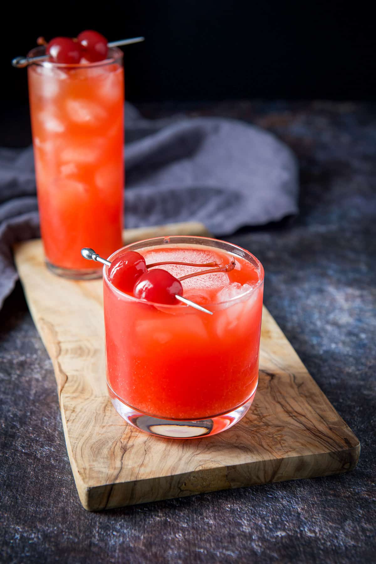A wooden board with two red cocktails garnished with cherries