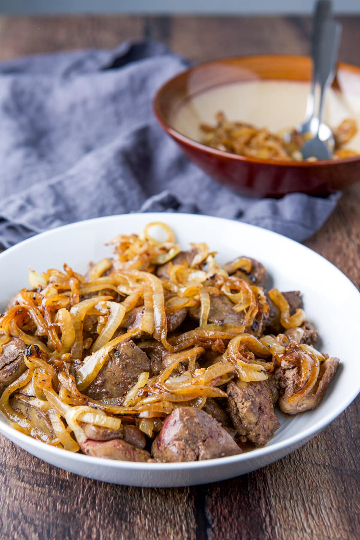 A wood table with a bowl of onions behind a white plate of livers and onions