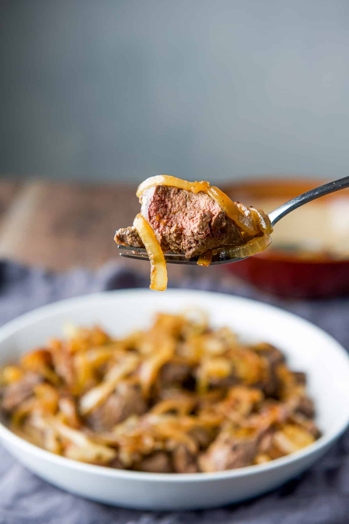 A fork with a bite taken out of a liver with onion on it over a plate of the livers
