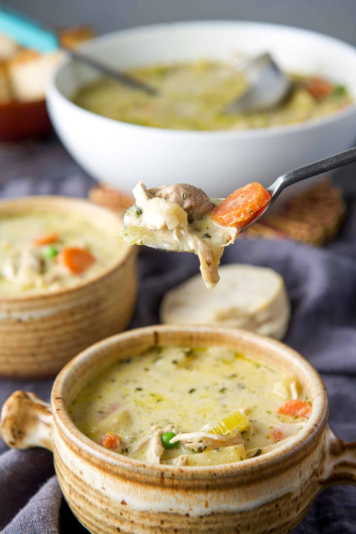 A spoonful of chicken soup held over a crock with another crock and serving bowl in the back
