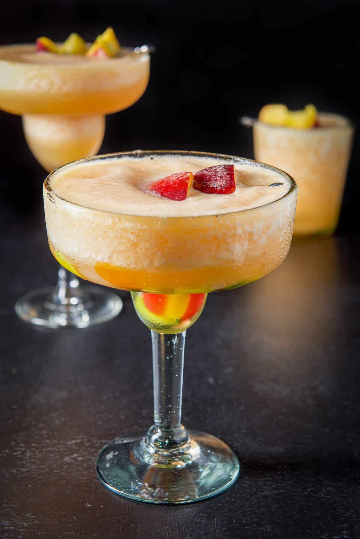 A colorful margarita glass with the peach margarita in it with two glasses behind it