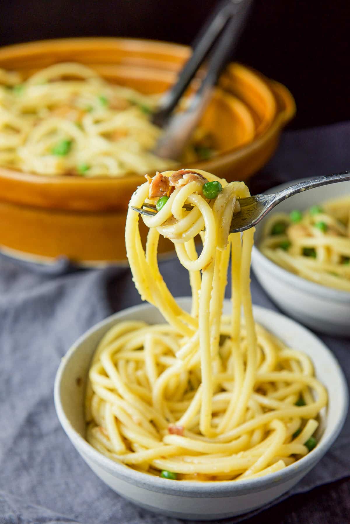 Forkful of carbonara held over the grey bowl with the brown serving bowl behind