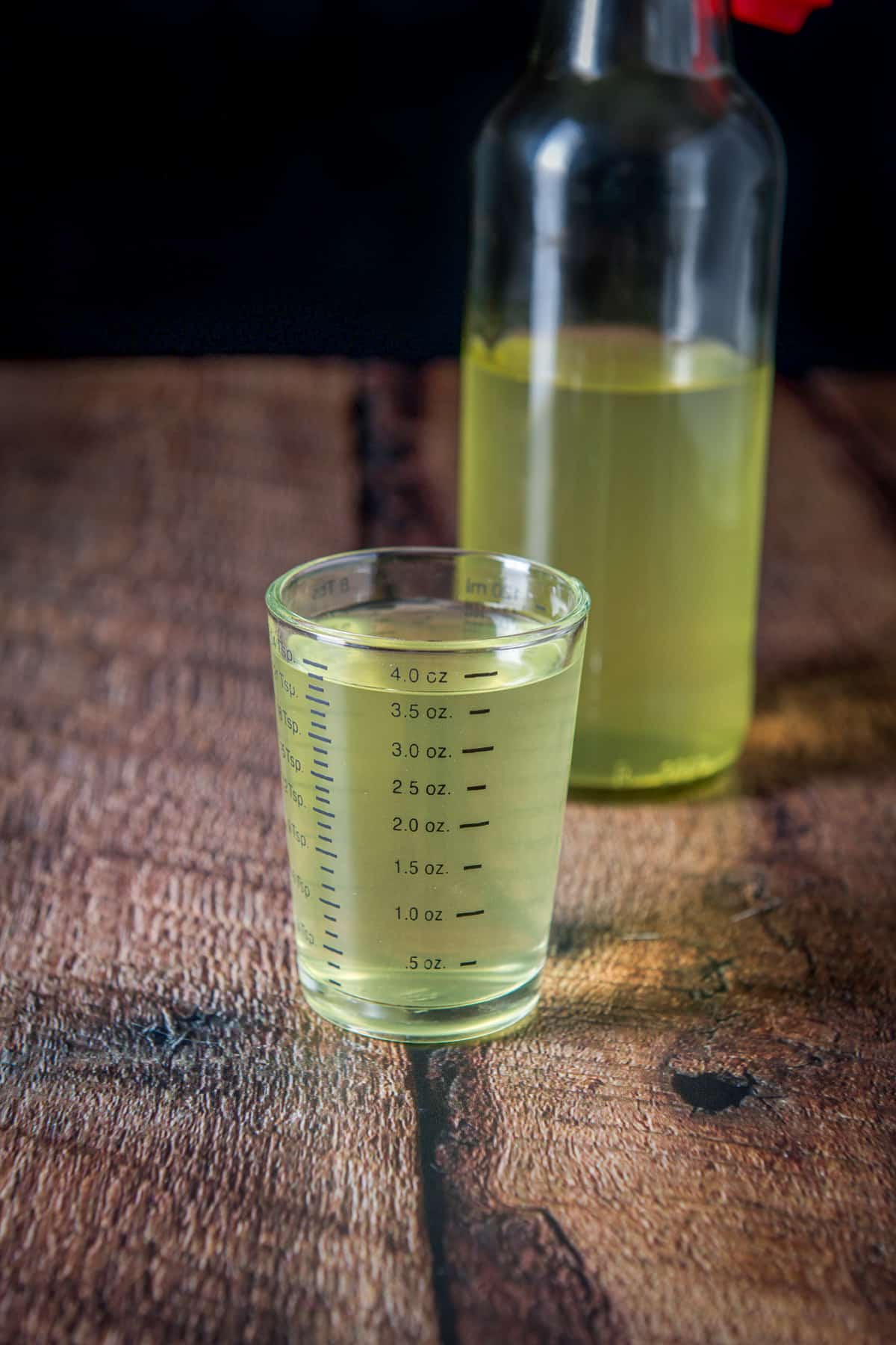 A measuring glass with the vodka in it with the bottle of lemon vodka in back