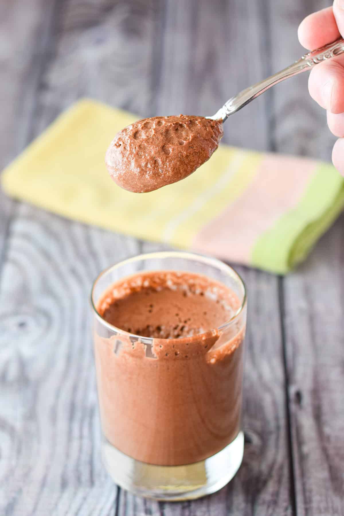 A hand holding a spoon with chocolate mousse on it held over the glass