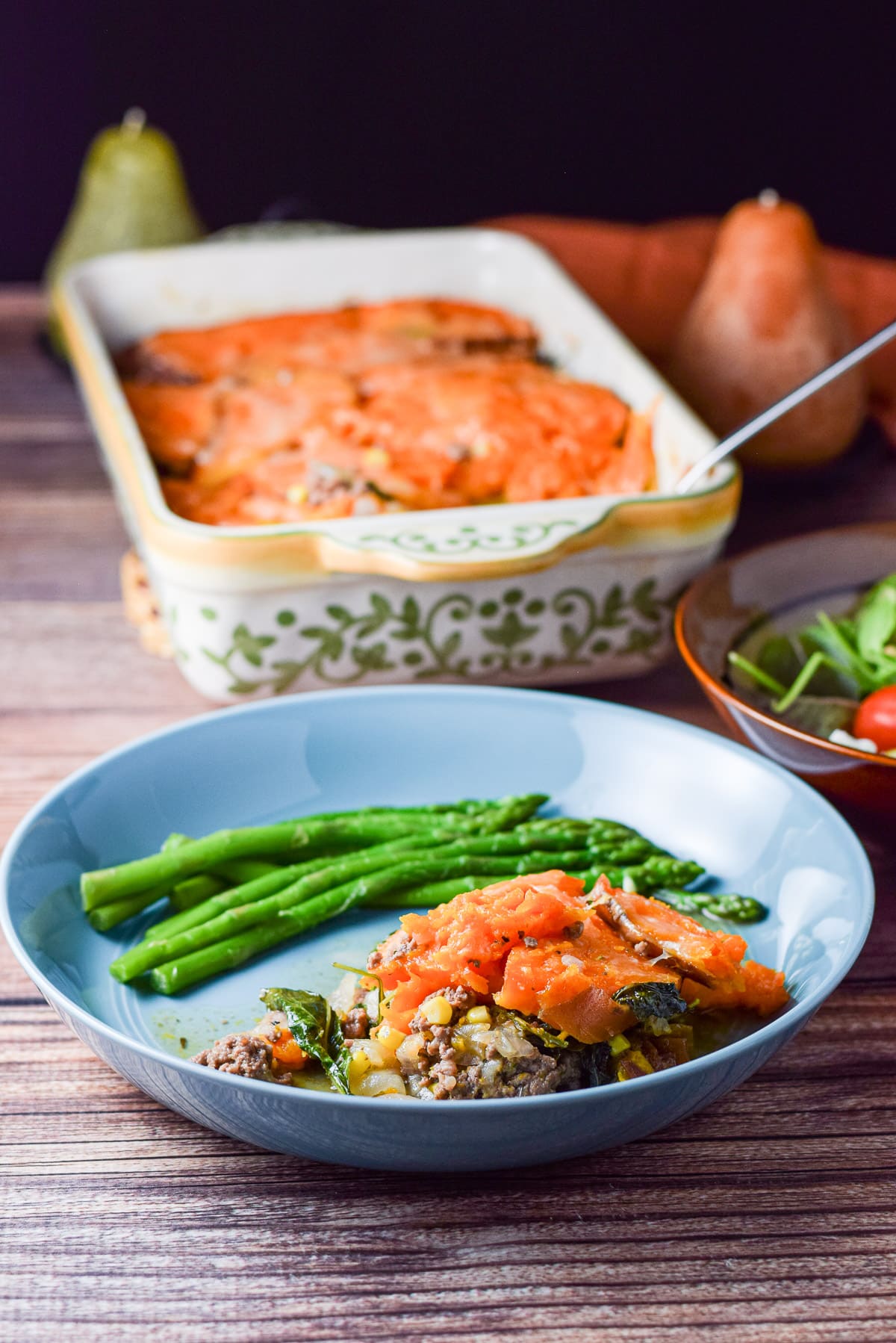 A casserole dish behind a deep plate with asparagus and the yam covered meat pie