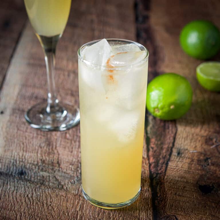 A tall glass with an amber cocktail with limes on the table - square