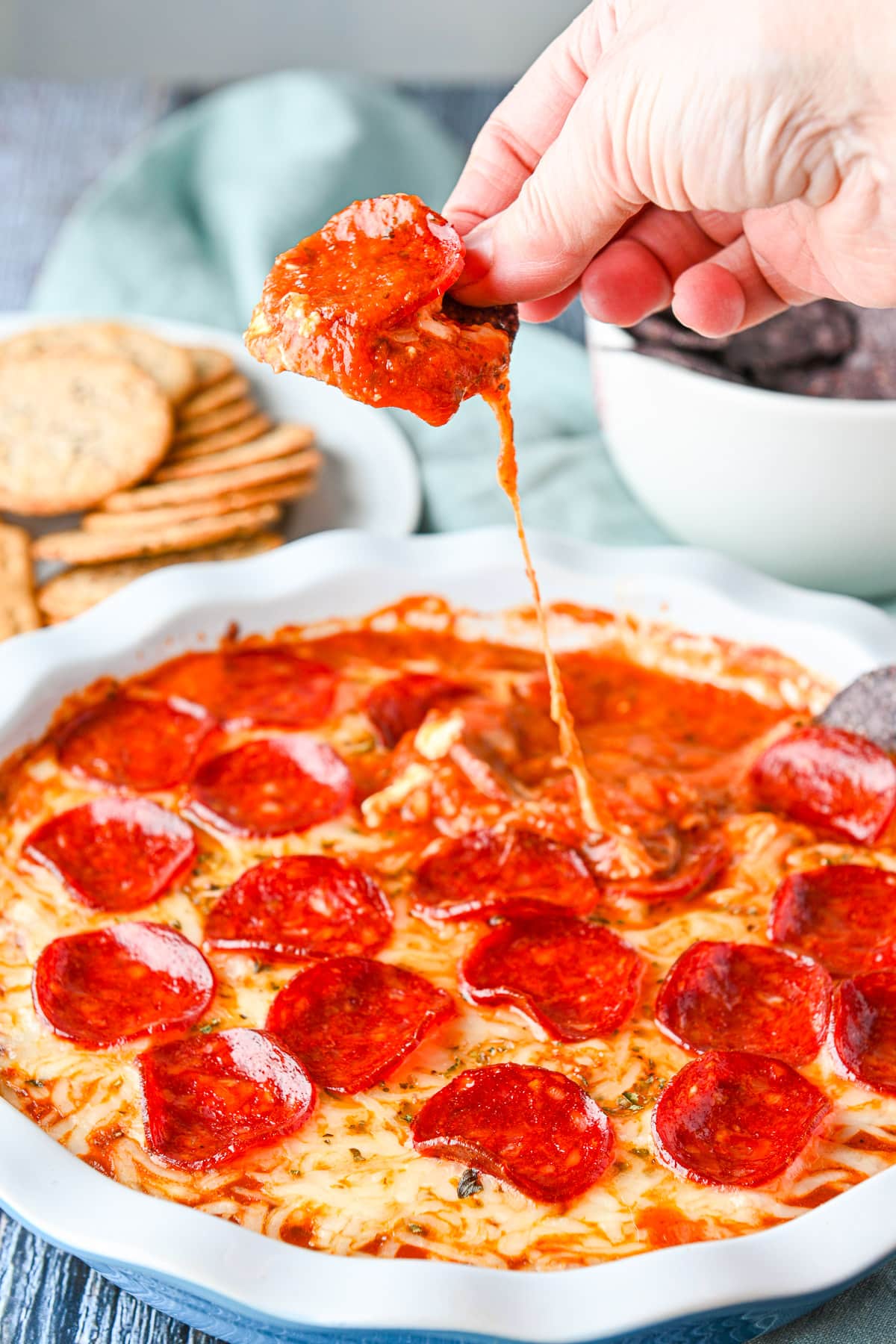 A hand holding a black corn chip with the pepperoni dip on it with a cheese string swinging down to the dip