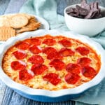 square photo of a pepperoni dip in a pie dish with chips and crackers in the back