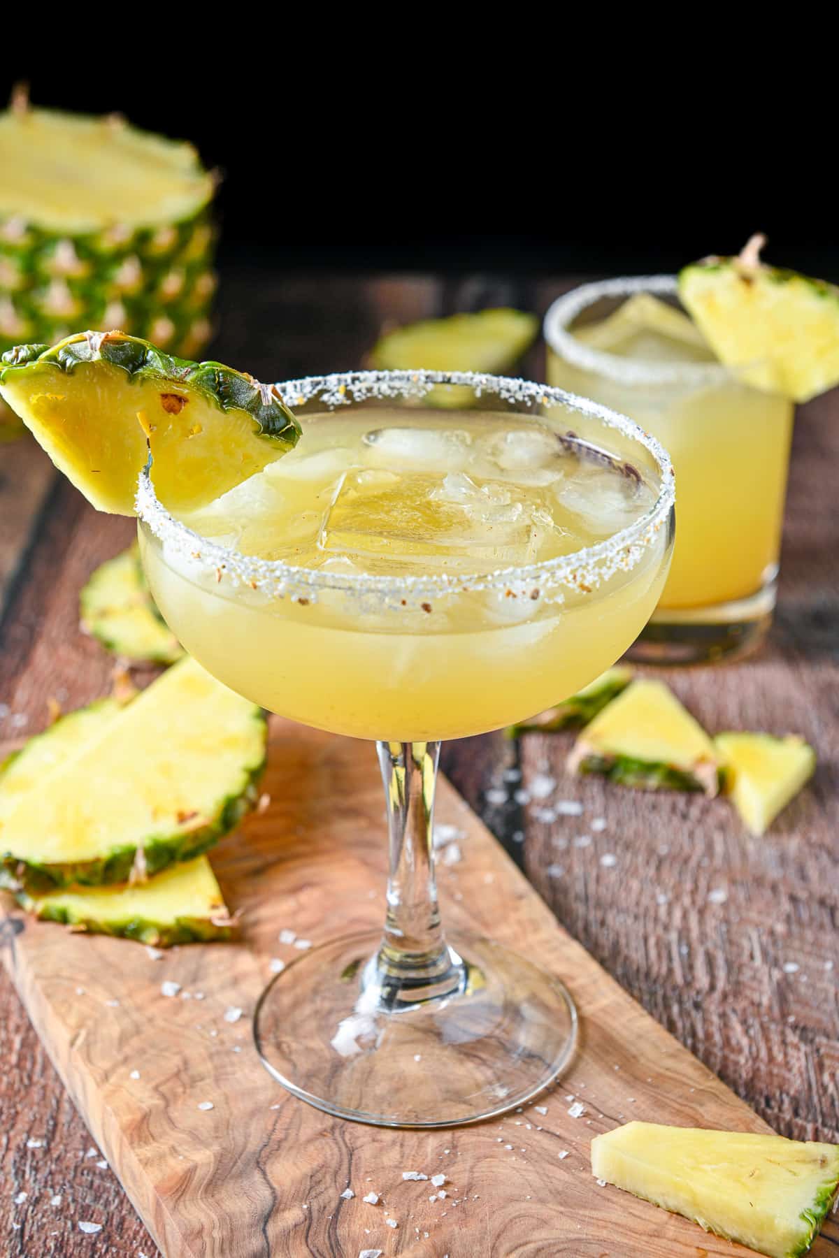Two margarita glasses on a table, one on a board and one in the back - both have salt on the rim and pineapple slices