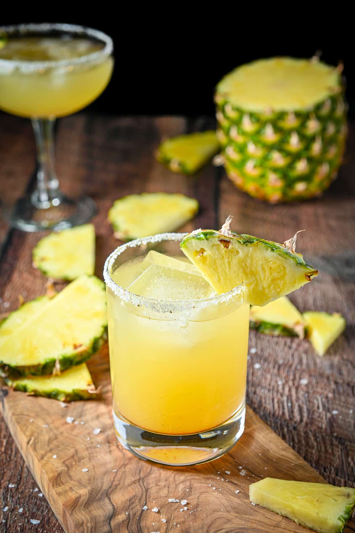 A double old fashioned glass with pineapple on the salted rim and the margarita in it. There are more slices of pineapple in the back