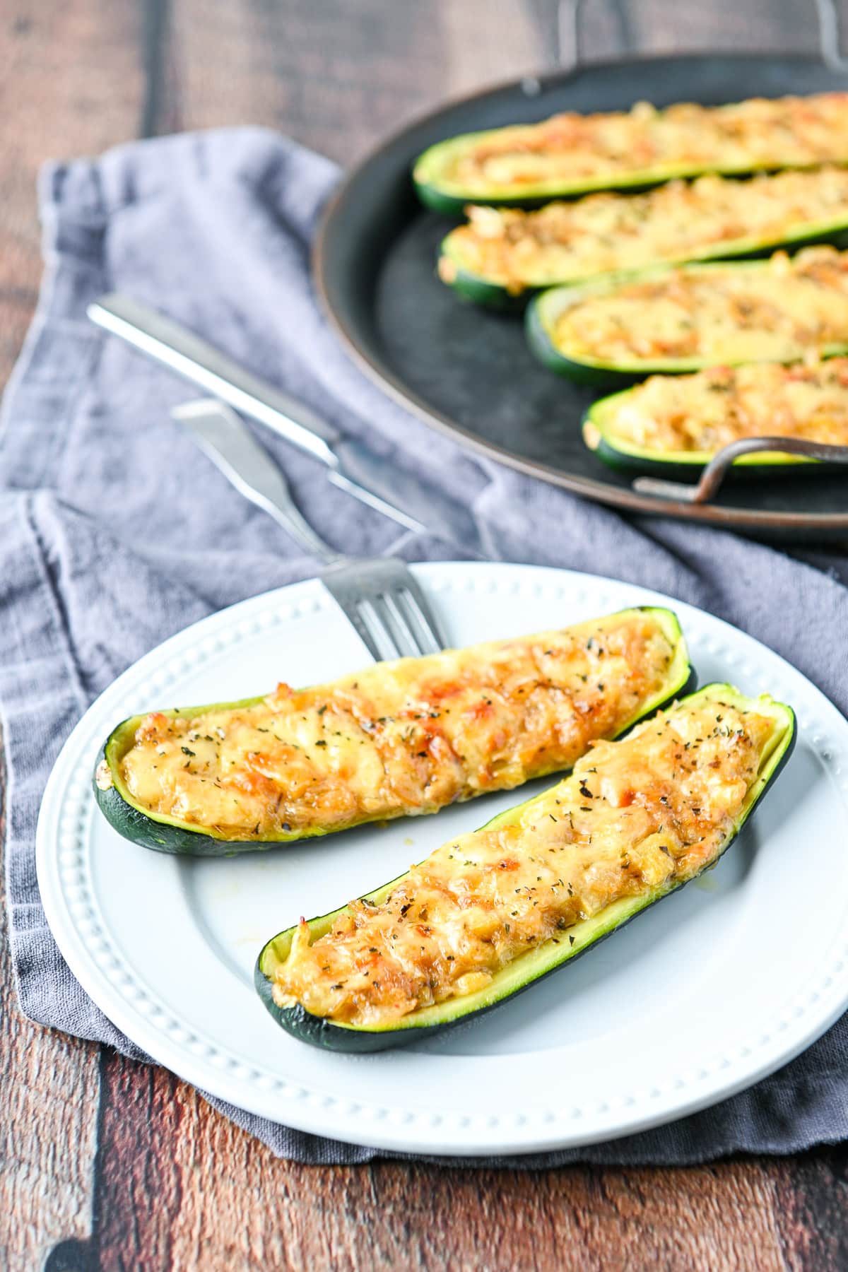 Two halves of zucchini on a white plate with a fork and knife and the tray of zucchini behind