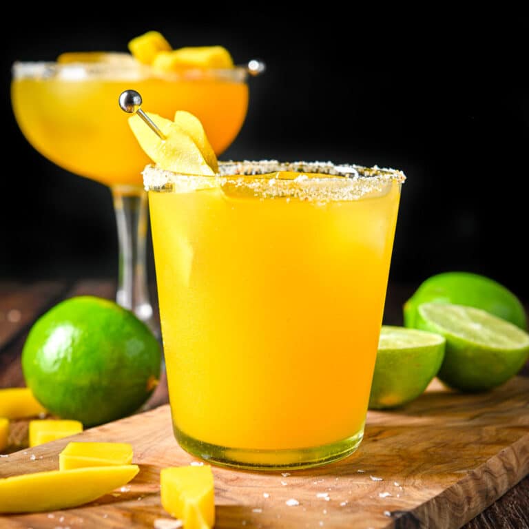 Square photo of two margaritas in vertical view with salt on the rims, and mango slices and limes on the table