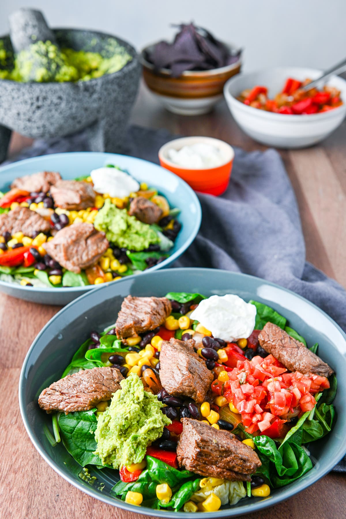 A table filled with burrito bowls with a molcajete filled with guacamole in the background with chips and condiments