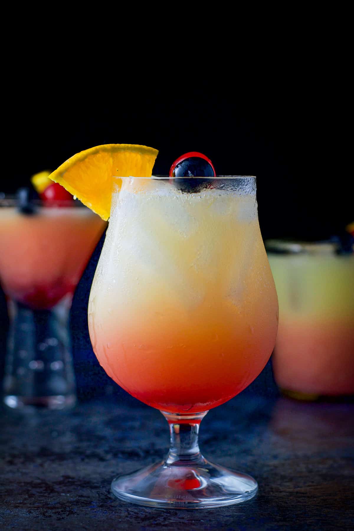 Vertical view of the tulip glass in front filled with the tequila sunrise. There are cherries and orange wedges as garnish