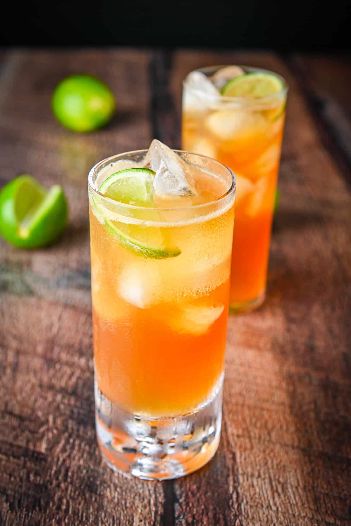 Two tall glasses with two dark and stormy cocktails in it along with limes on a table