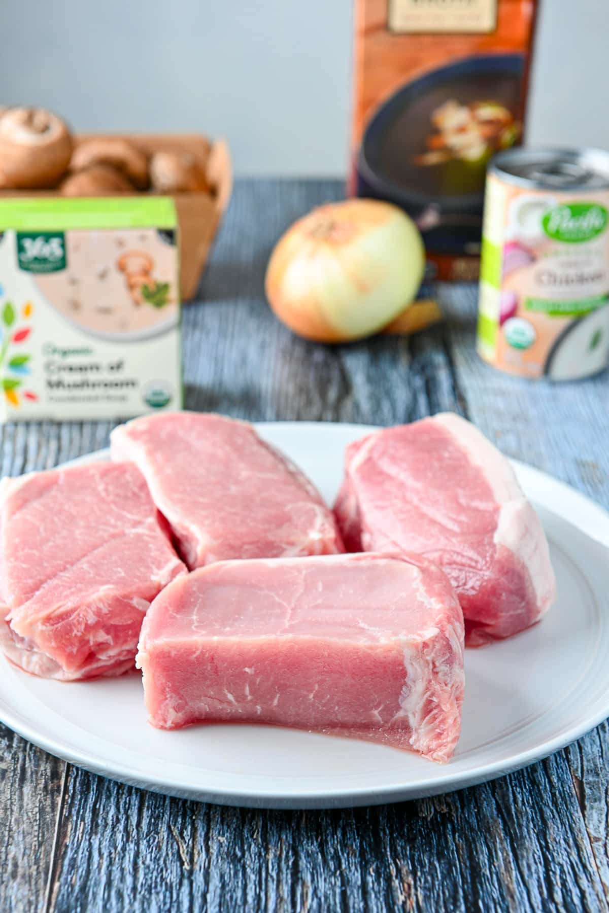 a white plate with four raw pork chops on it, condensed soup, onion, mushrooms in the back