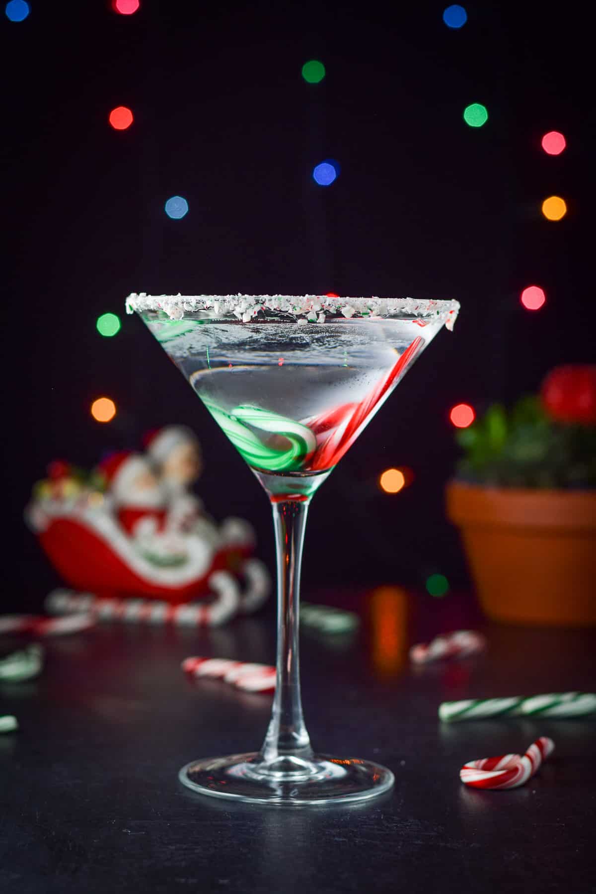 Vertical view of the mint cocktail with candy canes in the cocktail along with lights and holiday items in the back