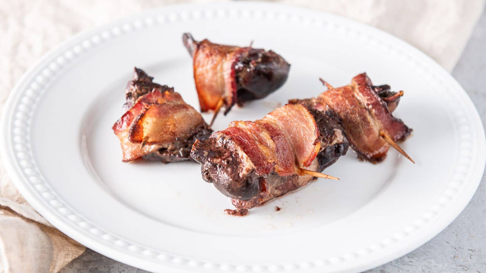a white beaded plate with four chicken livers wrapped with bacon and toothpicks through them