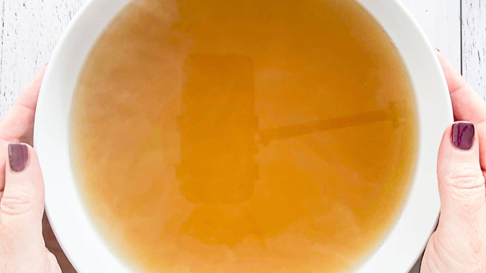 two hands holding a white bowl filled with chicken stock