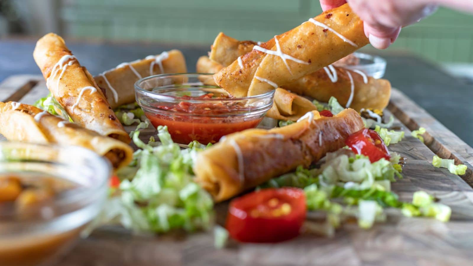 a hand holding a taquito in a bowl of salsa with more taquitos around it