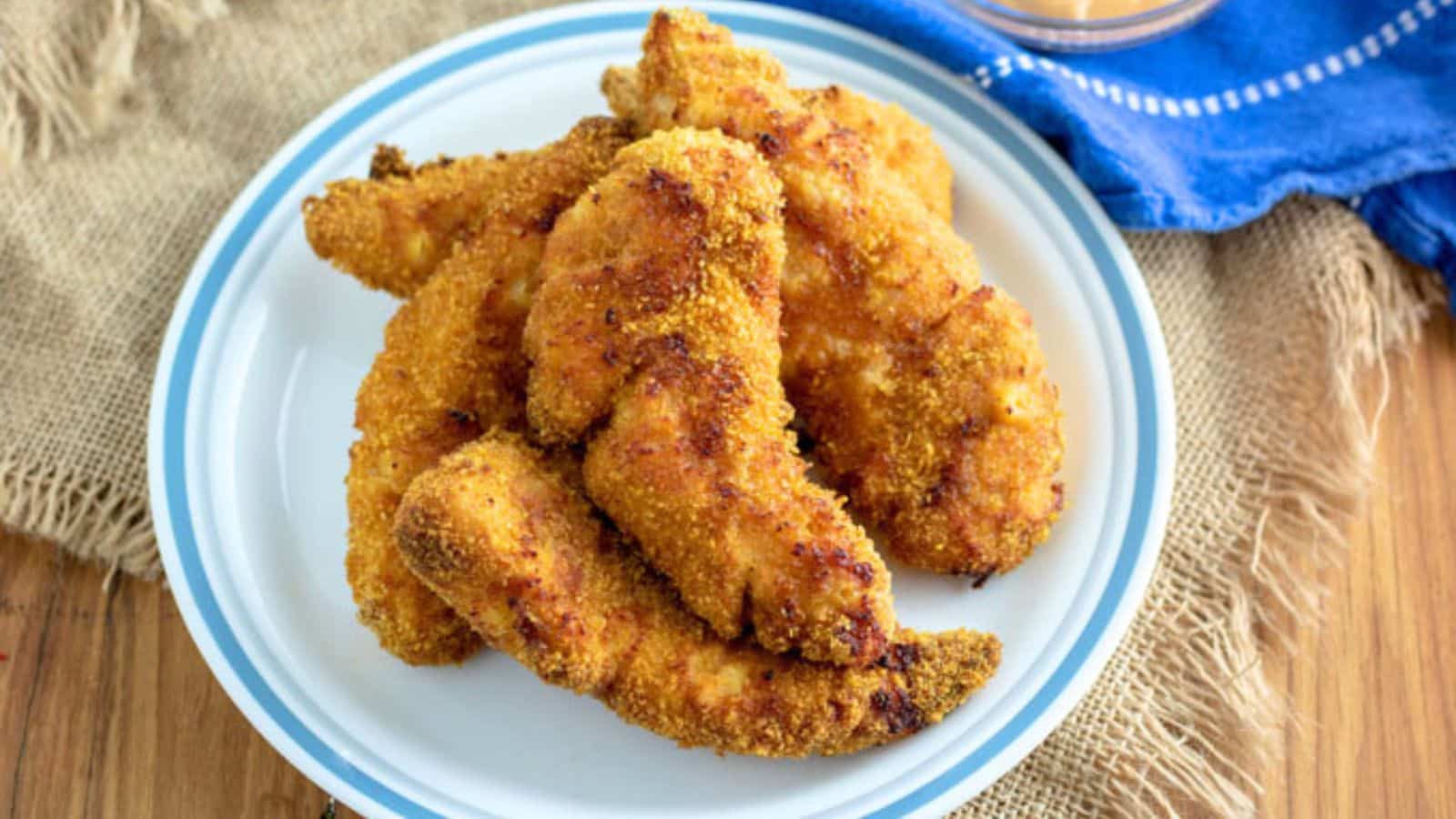 fried breaded chicken tenders on a white plate with burlap under the plate and a blue towel off to the side