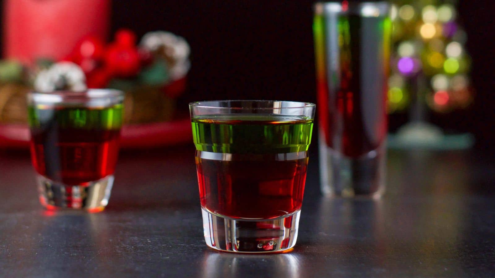 Three shot glasses filled with the red and green liquid in front of lights and a red candle