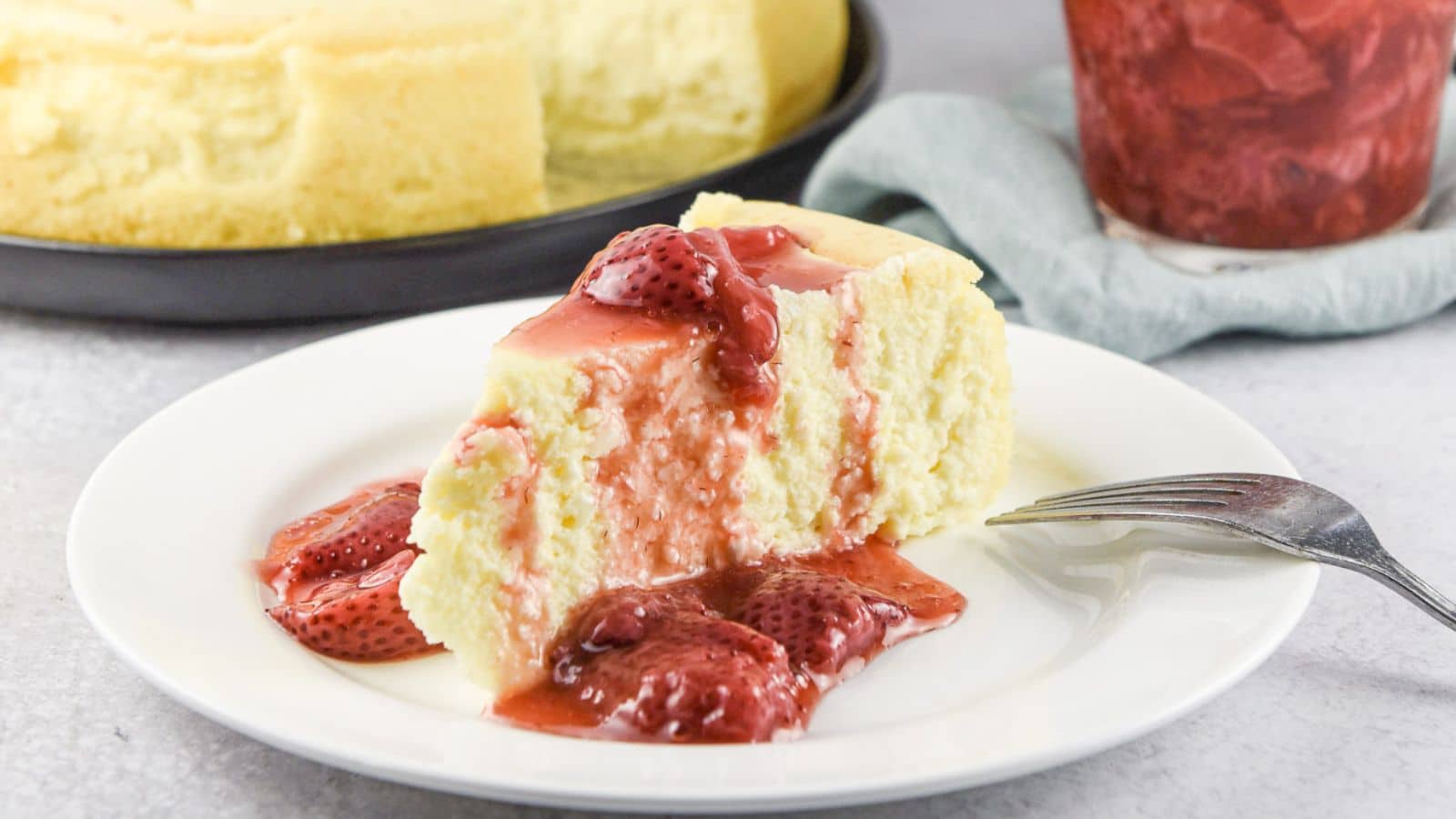 A white plate with a piece of cheesecake with strawberry sauce draped on it with the rest of the cheesecake in the back