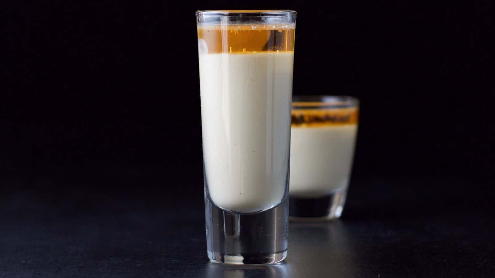 A tall glass in front of a short one filled with eggnog and a light amber liquid on top