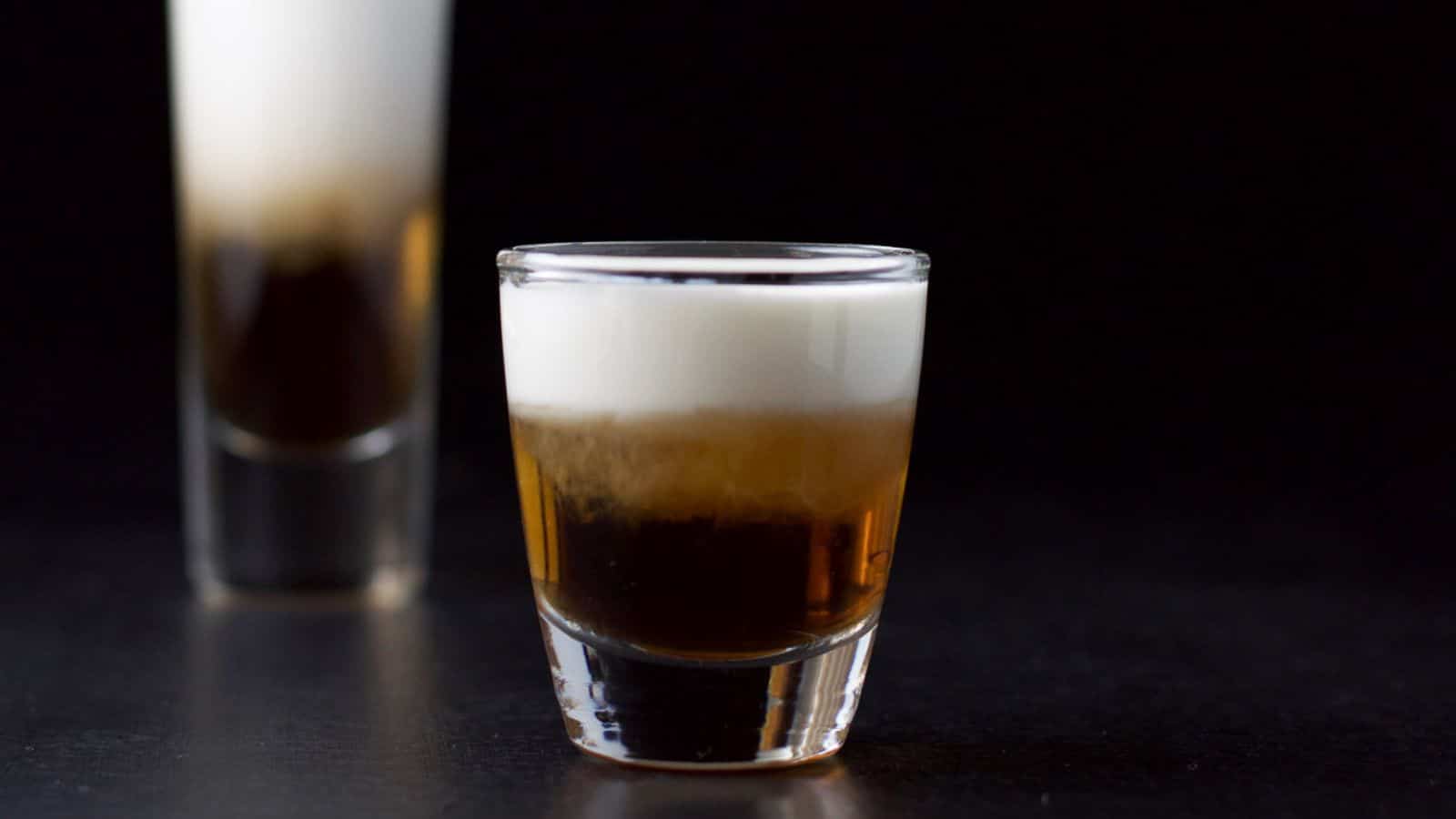 a short glass layered with an amber and white liquid in front of another taller glass