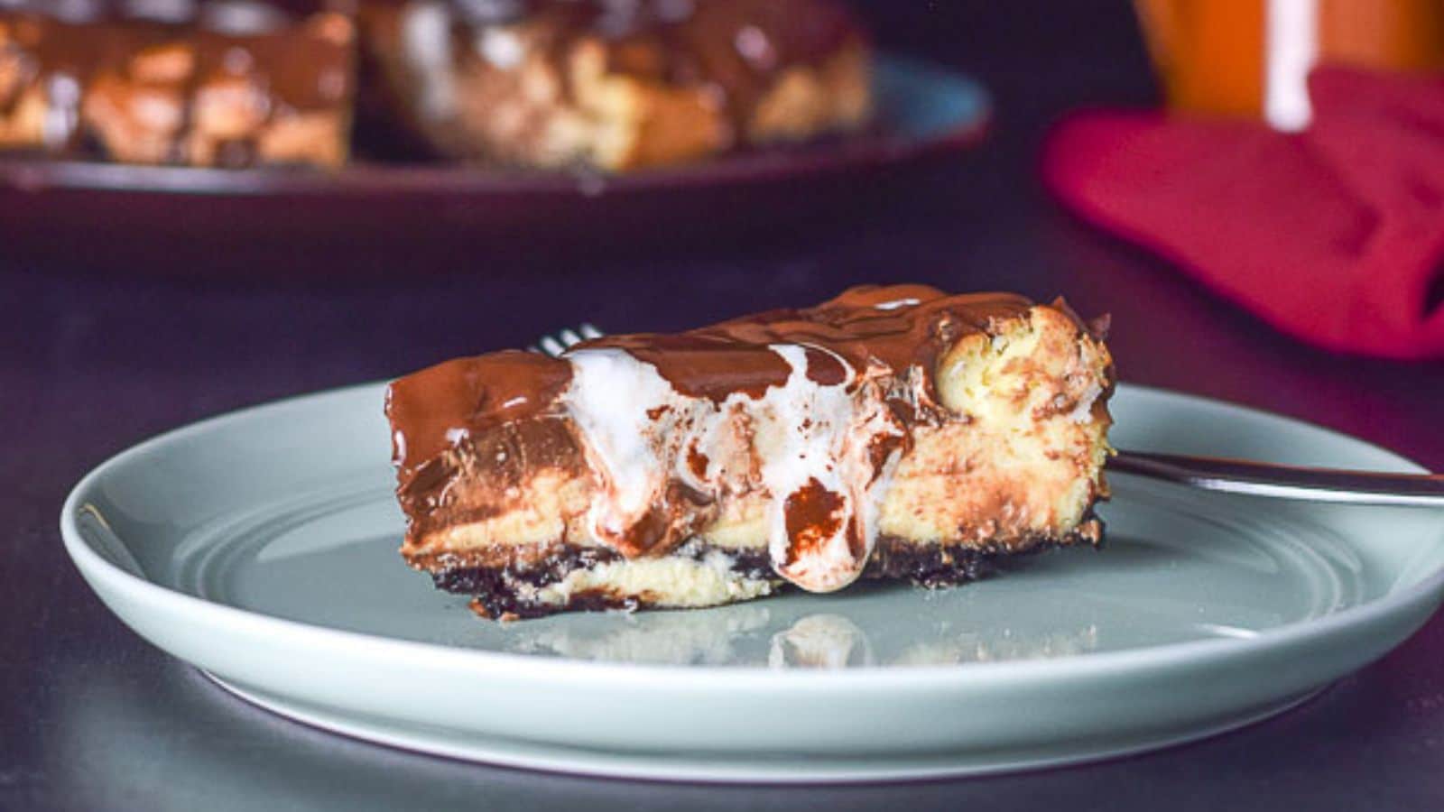 A slice of cheesecake on a green plate with marshmallow and chocolate with the cake in the back