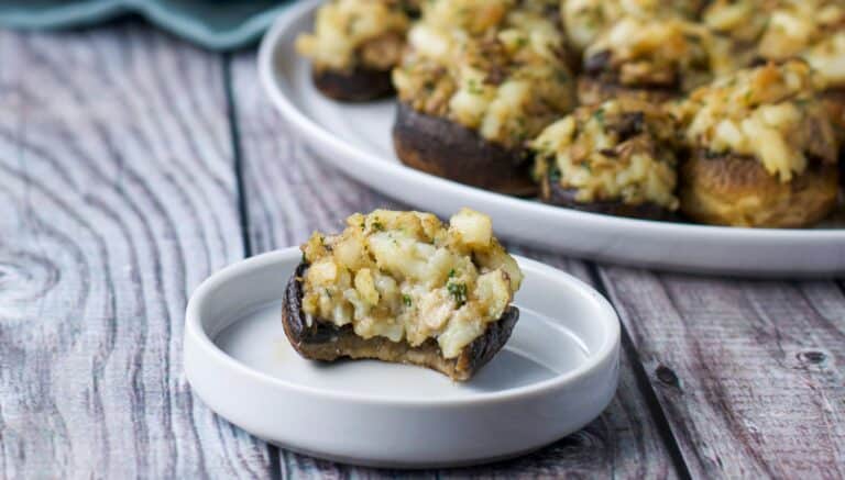 A small plate with a stuffed mushroom on it with a bite taken out in front of a plate of mushrooms