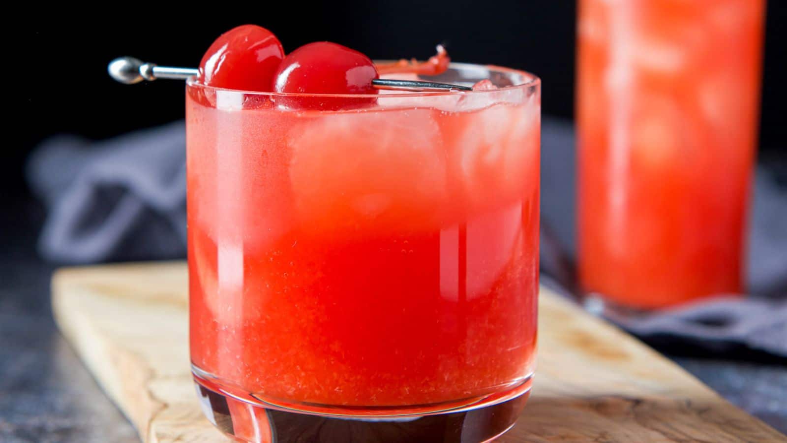 a glass on a board with the red cocktail with two cherries on a pick as garnish