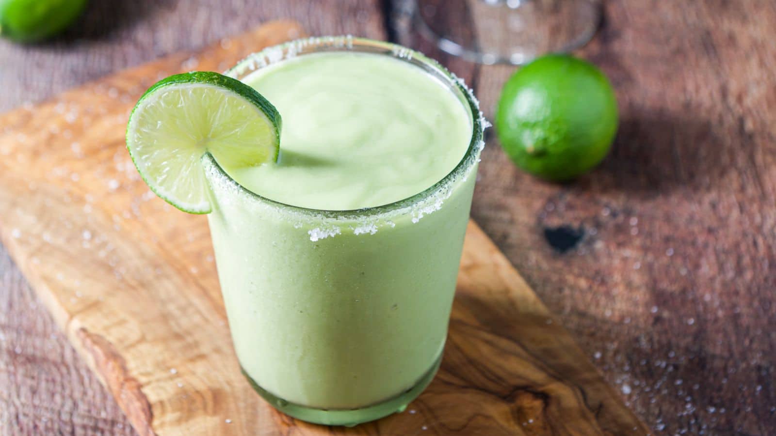 a glass with salt on the rim and a lime wheel filled with a thick green drink along with a lime on the table