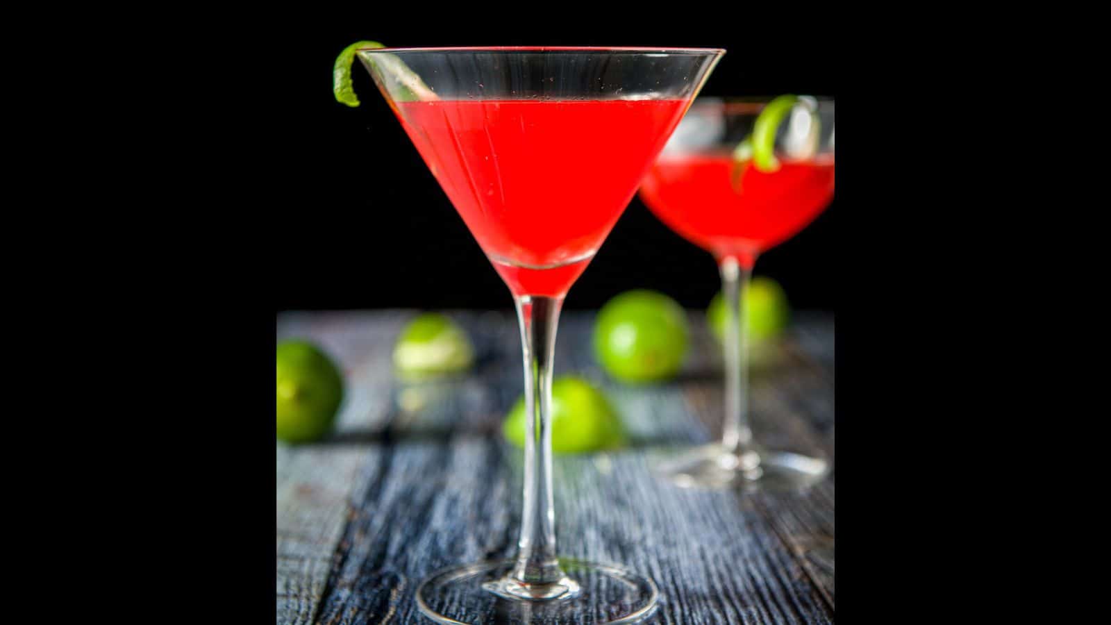 Two glasses filled with the Charlie Chaplin cocktail with lime twists as garnish and more limes on the table