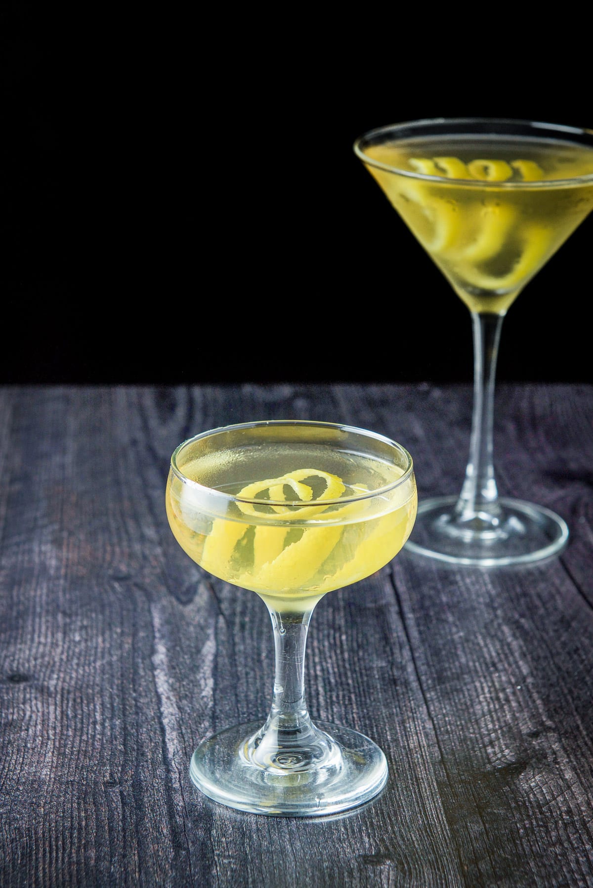 Coupe glass in front of the classic glass. The lemon twist is prominent in the cocktail