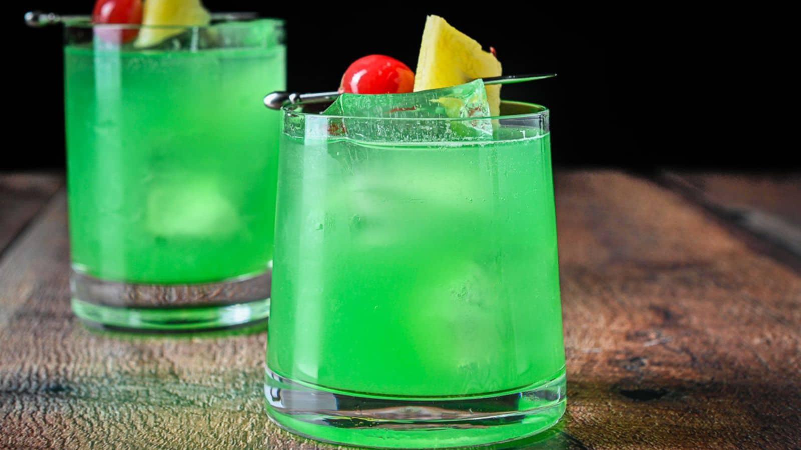 two glasses filled with the green cockatil with pineapple chunks and cherries as garnish