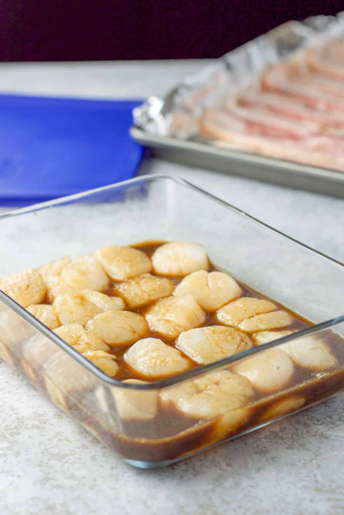 Scallops marinating in a glass dish and bacon on a foil lined pan