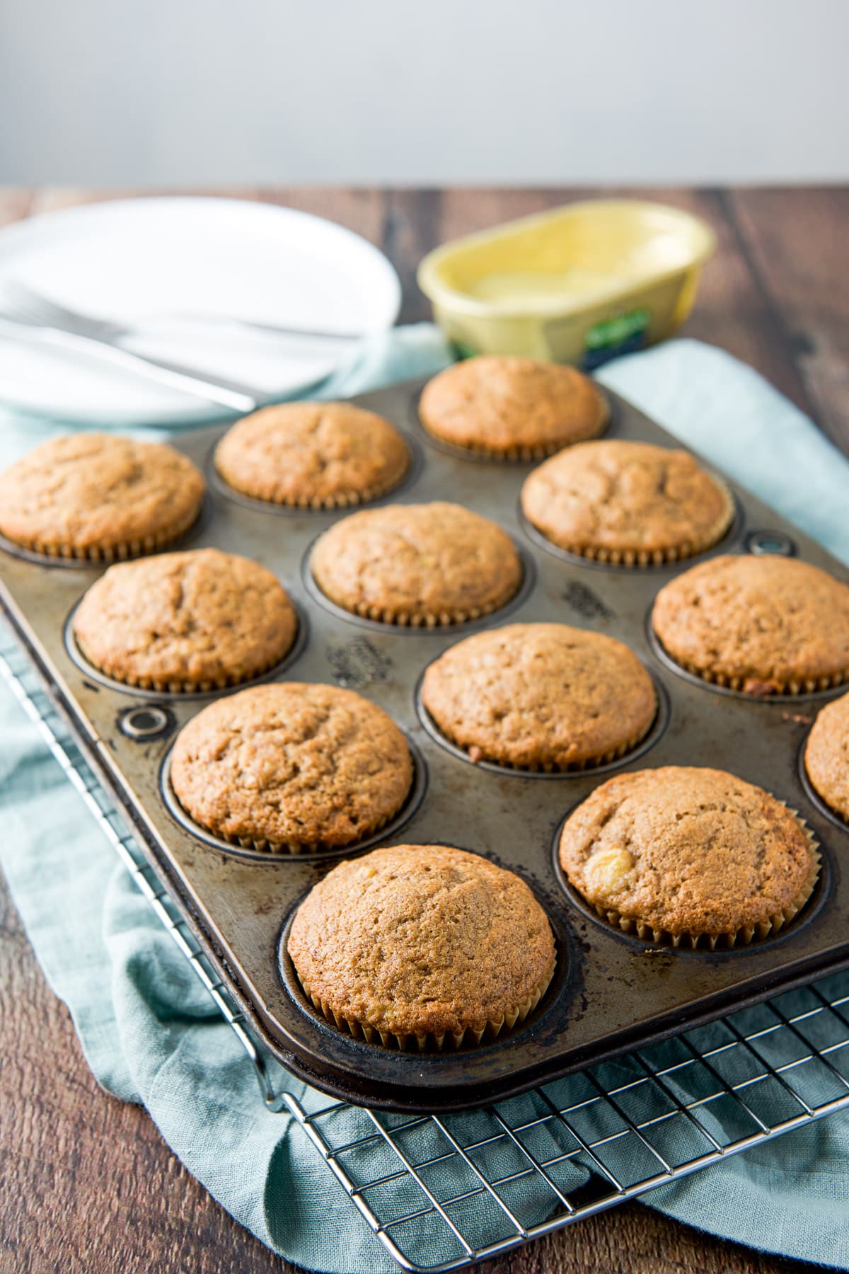 a muffin pan with brown muffins in it on a wire rack, with plates in the back and butter