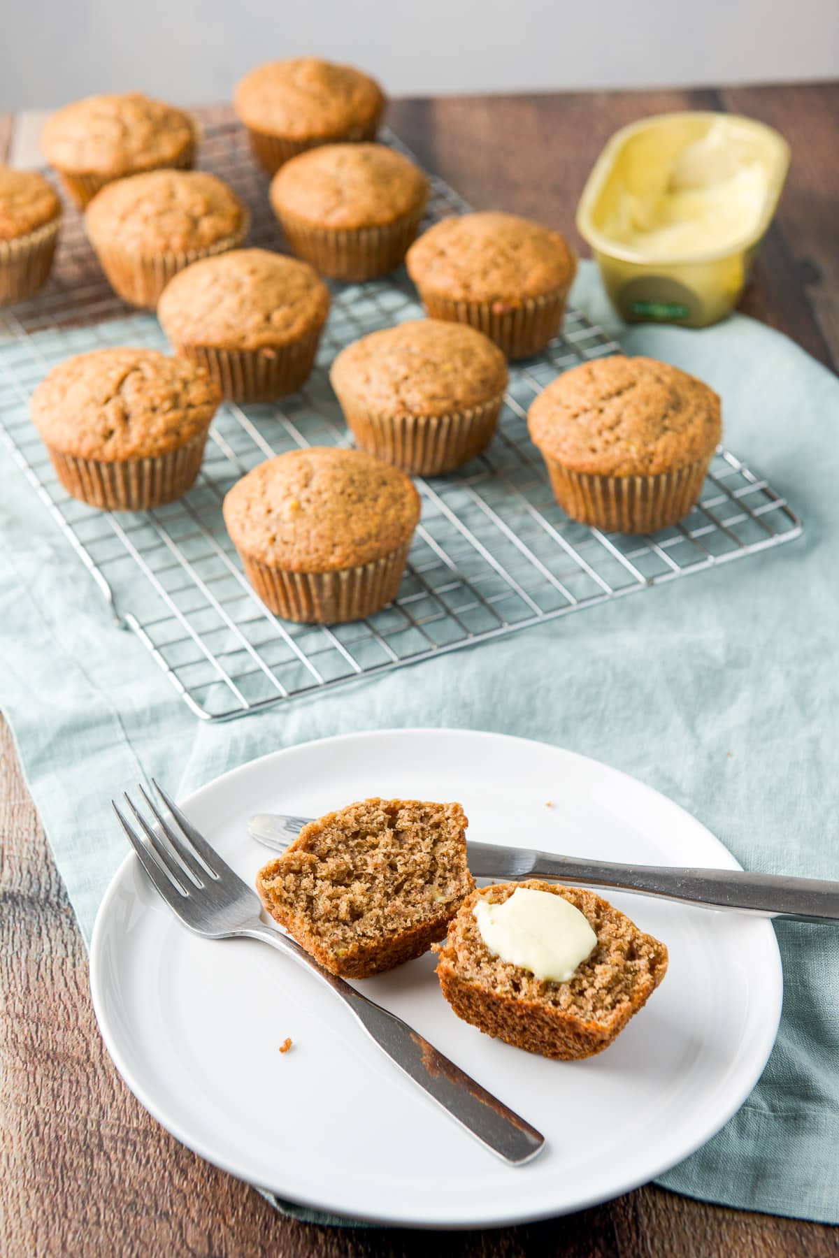a white plate with a muffin cut in half with butter on one side. There are more muffins in the background along with butter