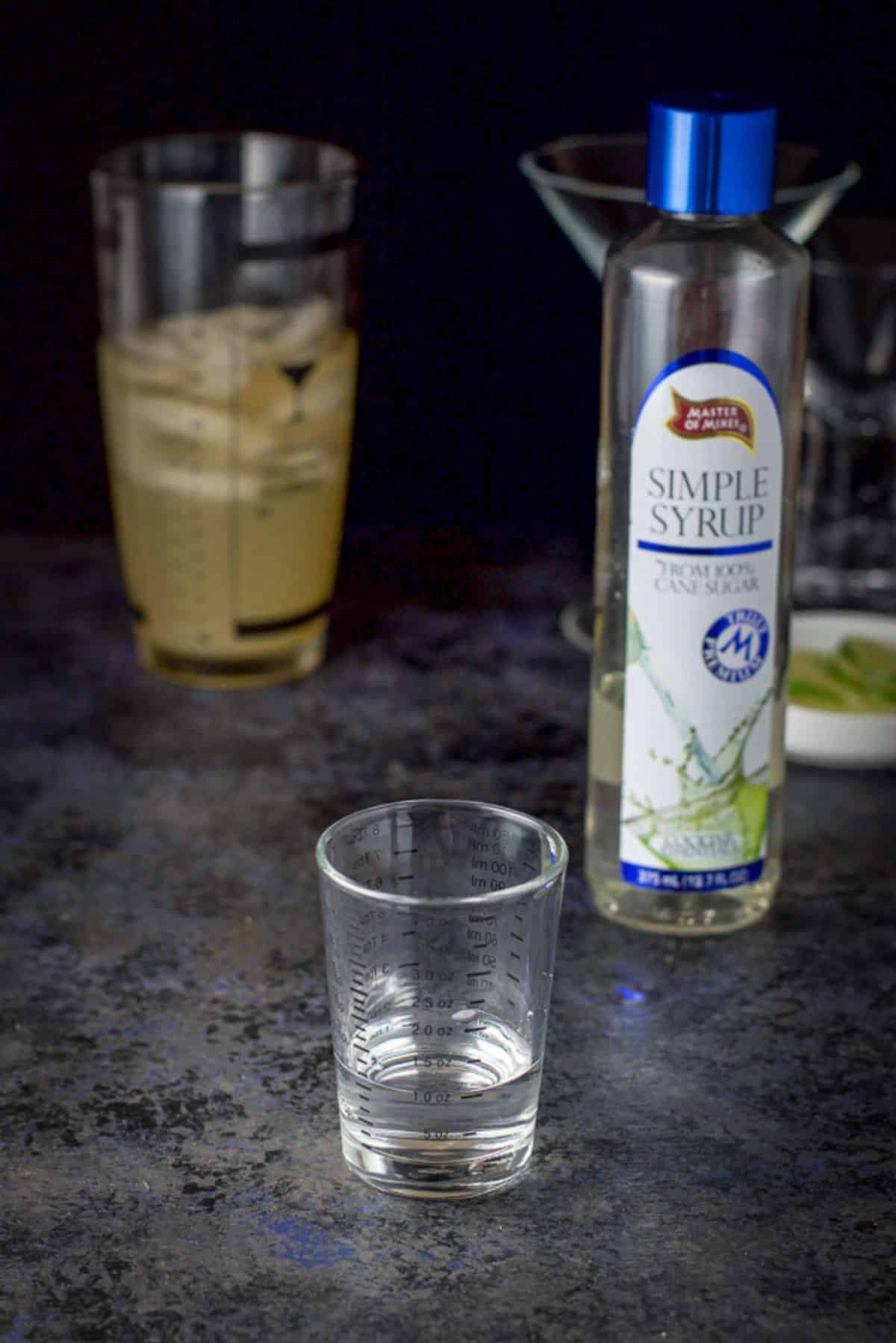 Simple syrup measured out with the bottle in the background and the shaker and glasses in the back as well
