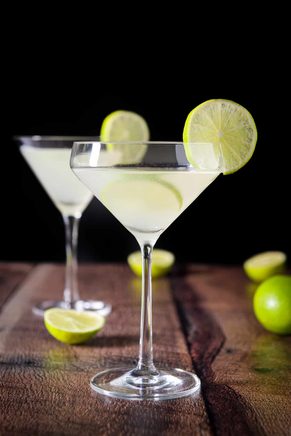 Vertical view two glasses filled with the kamikaze, with limes on the table and lime wheels on the side of the glasses