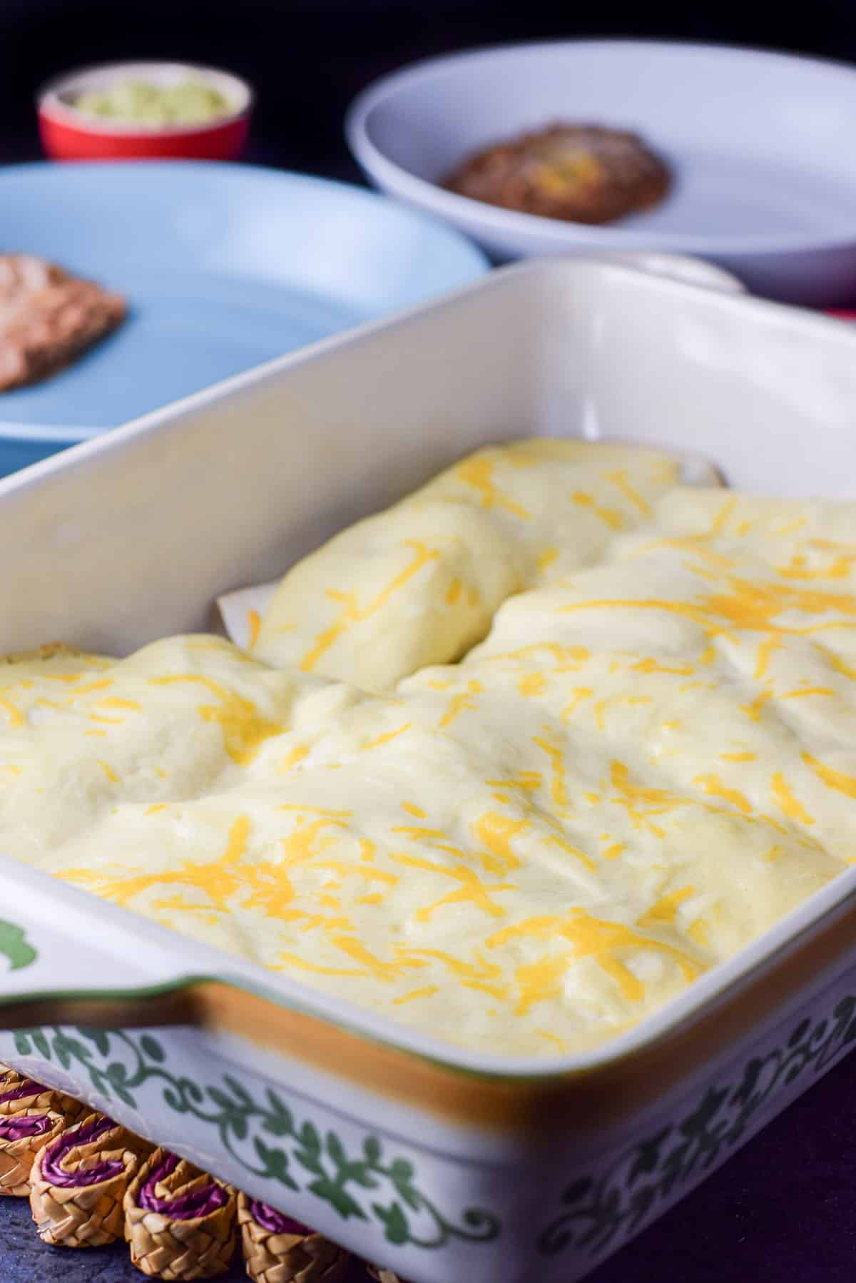 A 13 x 9 ceramic pan with baked enchiladas with sauce on top and plates with beans in the background