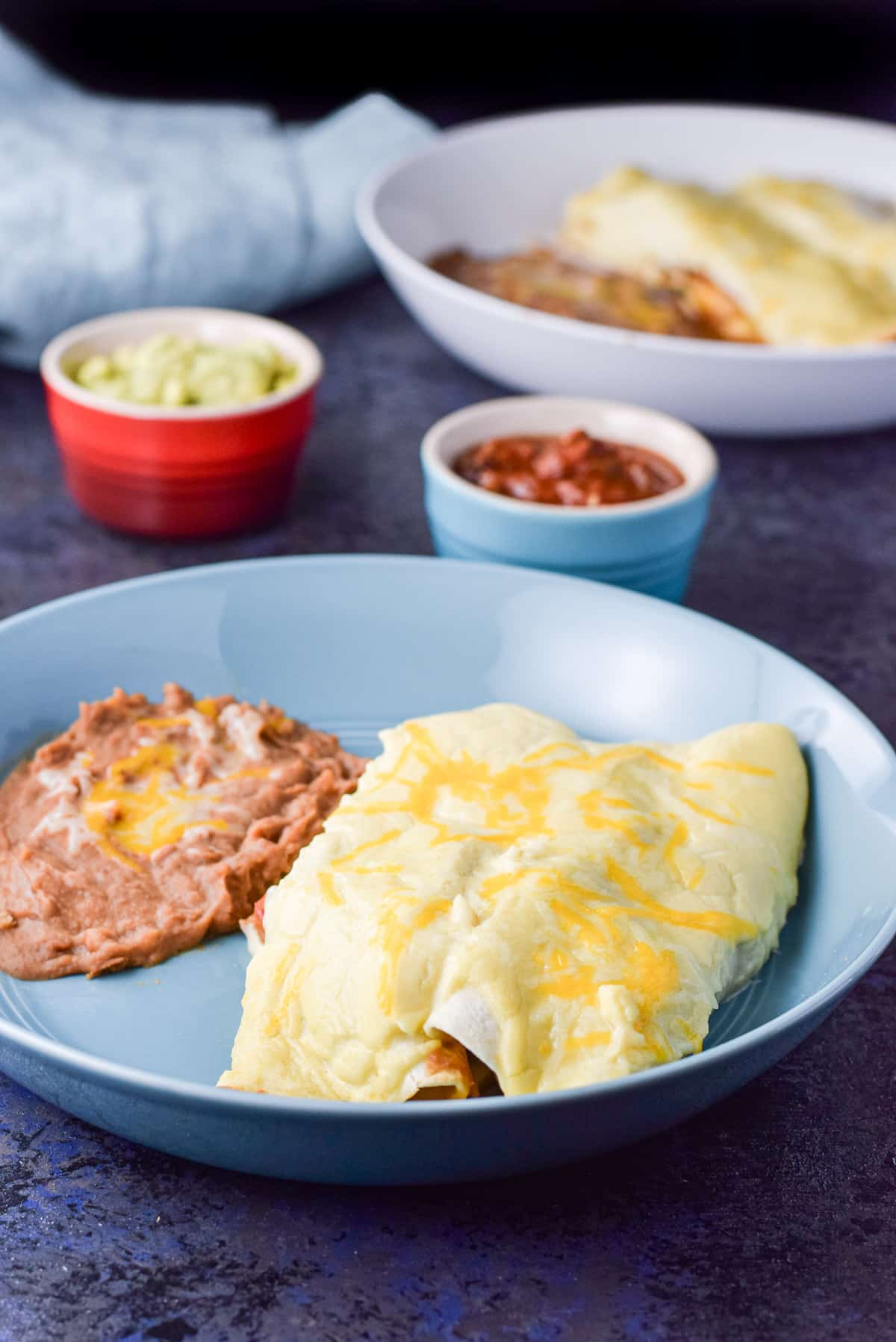 Two shrimp enchiladas on a plate with beans with guac and salsa along with another plate of the meal