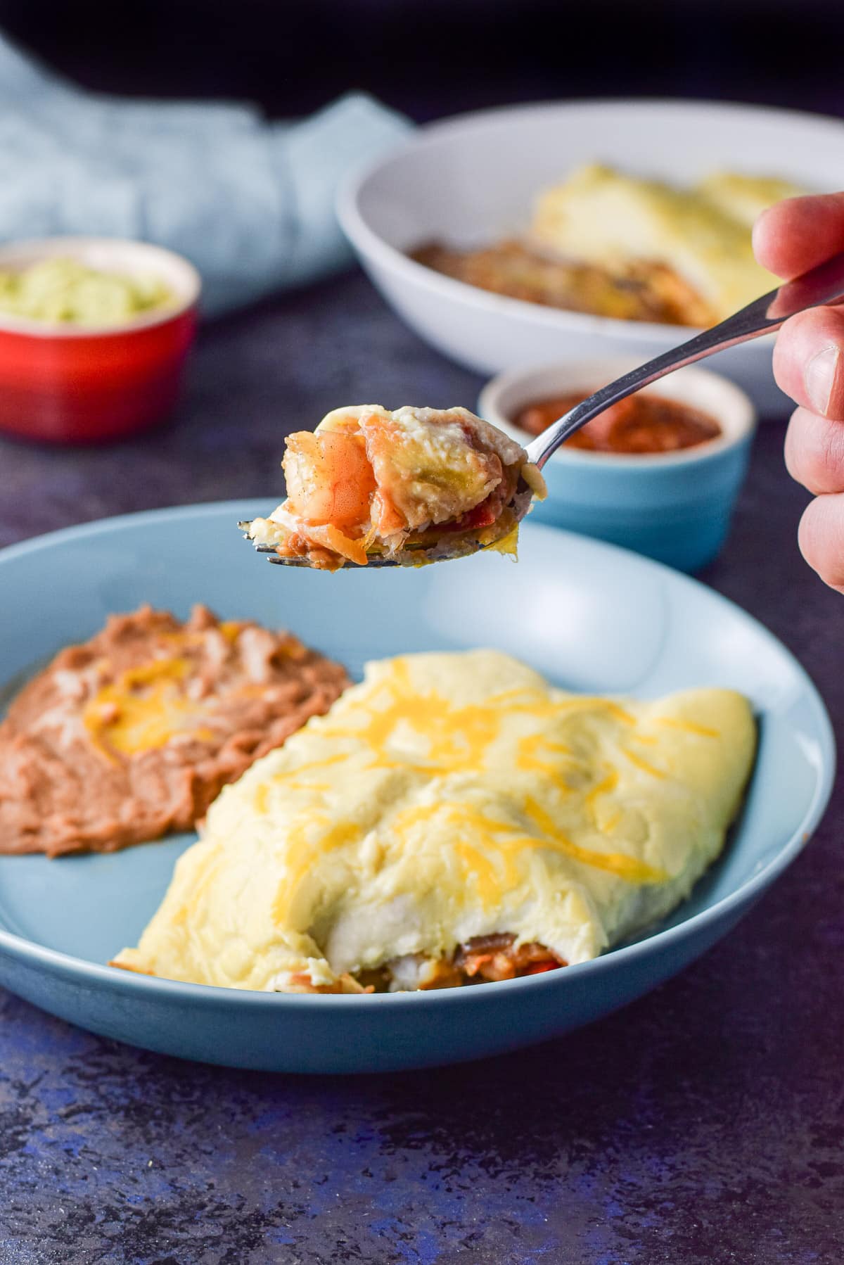 A hand holding a fork with shrimp in an tortilla held over a plate with the enchiladas and beans
