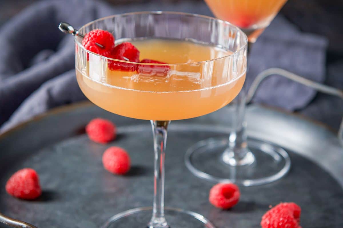 A coupe glass with an amber cocktail with raspberries as garnish