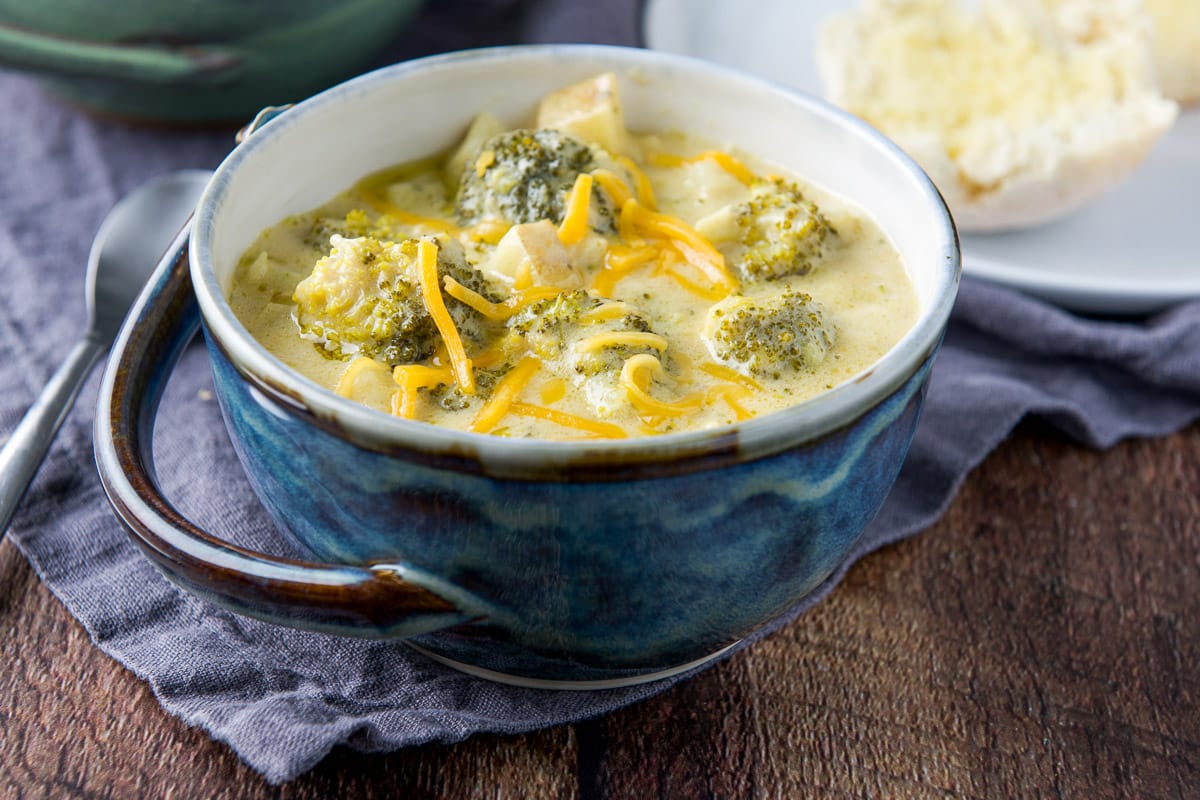 A blue bowl with a handle filled with broccoli soup with shredded cheddar on top and a roll in the back