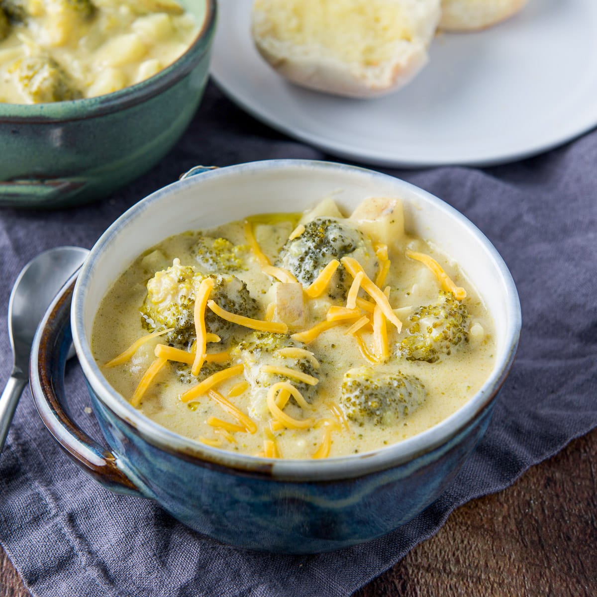 A blue bowl filled with the broccoli soup with cheddar on top. There is another bowl behind it and a roll on a plate