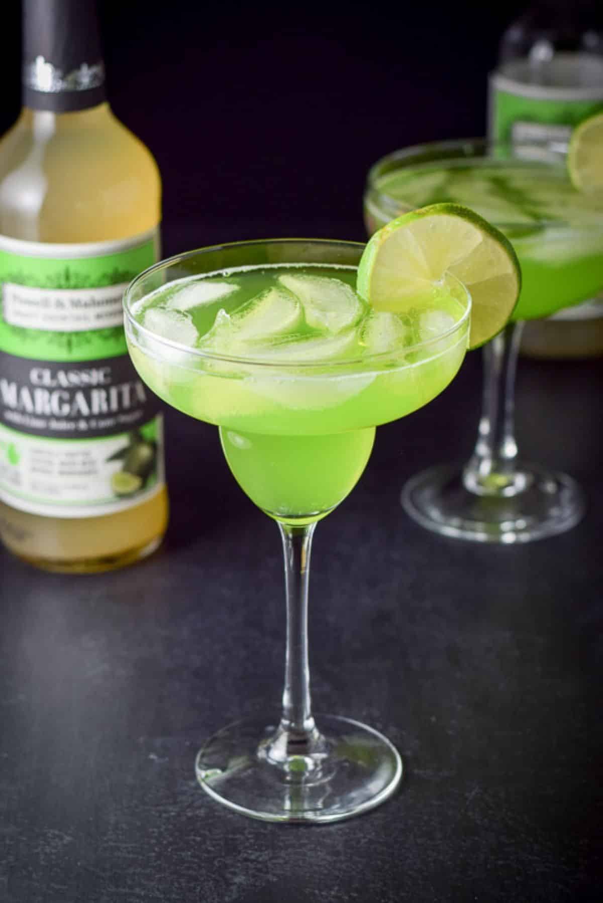 Two margarita glasses filled with the melon marg with the bottle of sweet and sour in the background