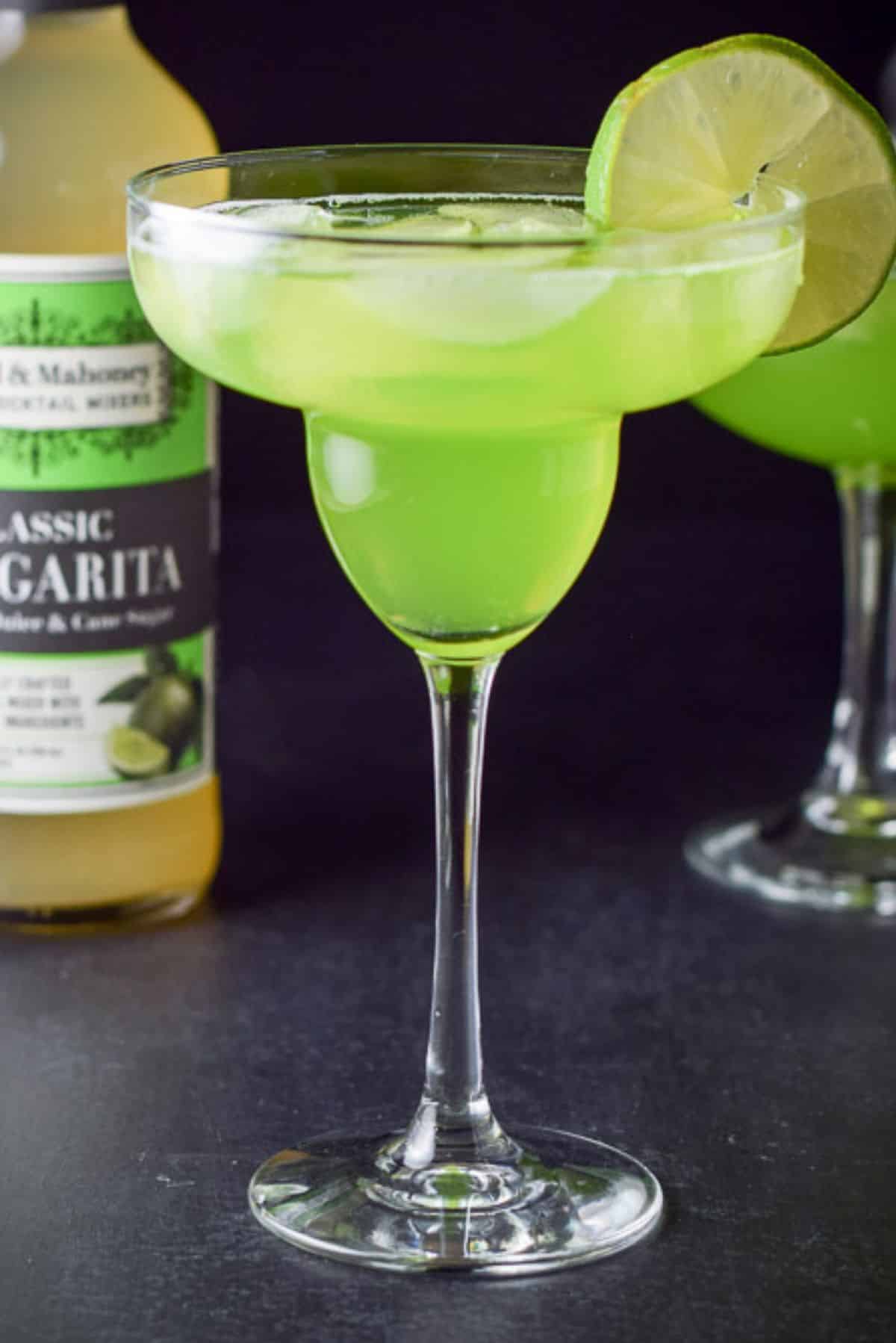 Vertical views of the margaritas with lime wheels on the side of the glasses and the bottle of sweet and sour in the background