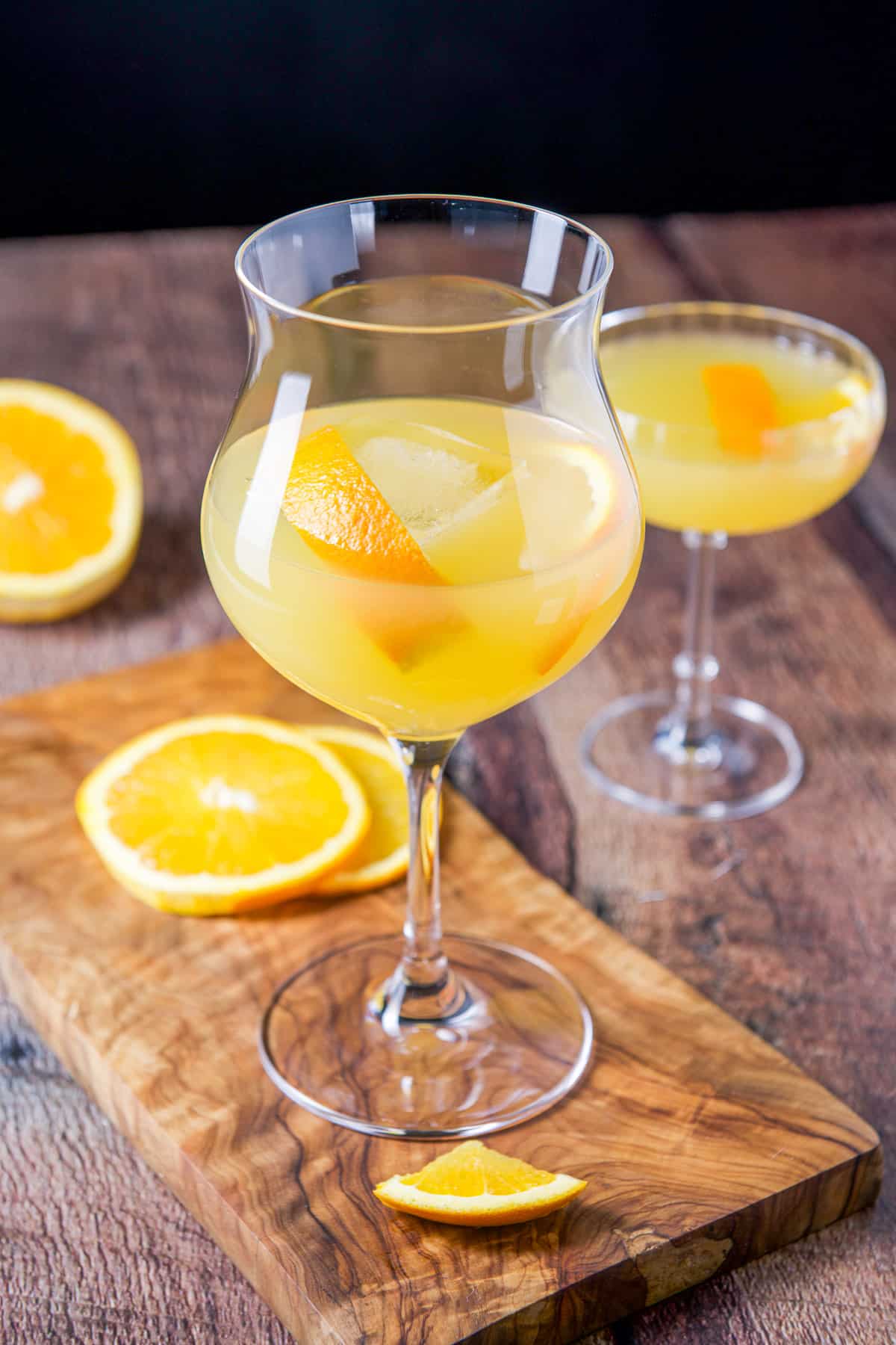 A tulip shaped glass and coupe glass filled with the paradise cocktail with orange twists as garnish with more orange slices on the table